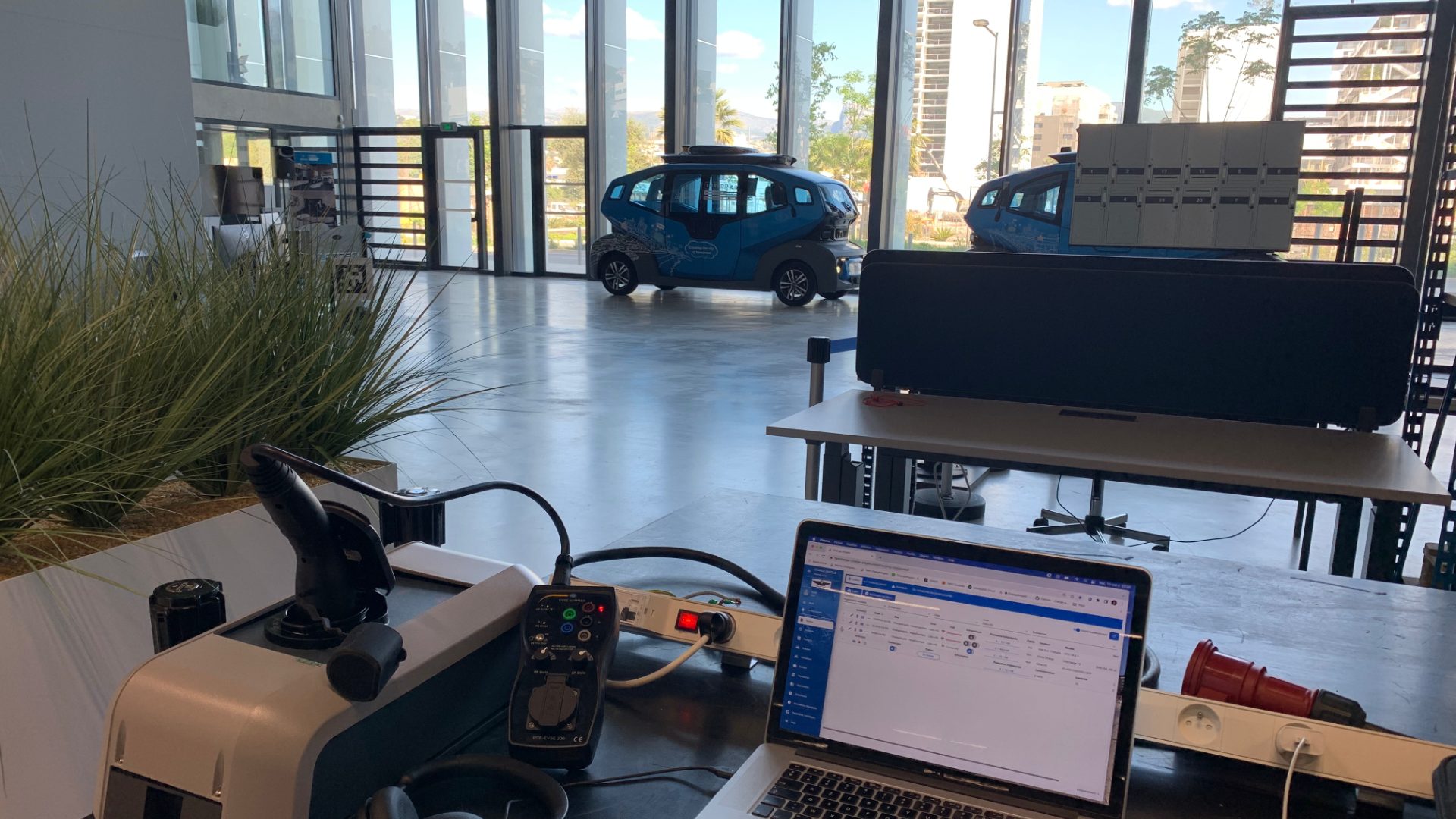 ChargeAngels test bench at IMREDD: an EV charging connector, an EVSE tester and a laptop showing the ChargeAngels dashboard, with a blue autonomous shuttle in the hall behind