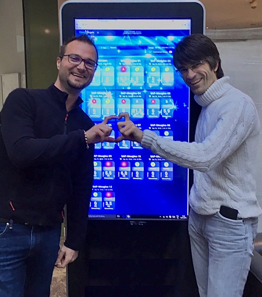 Two members of the ChargeAngels team making a heart with their hands in front of the ChargeAngels dashboard on a large touch screen